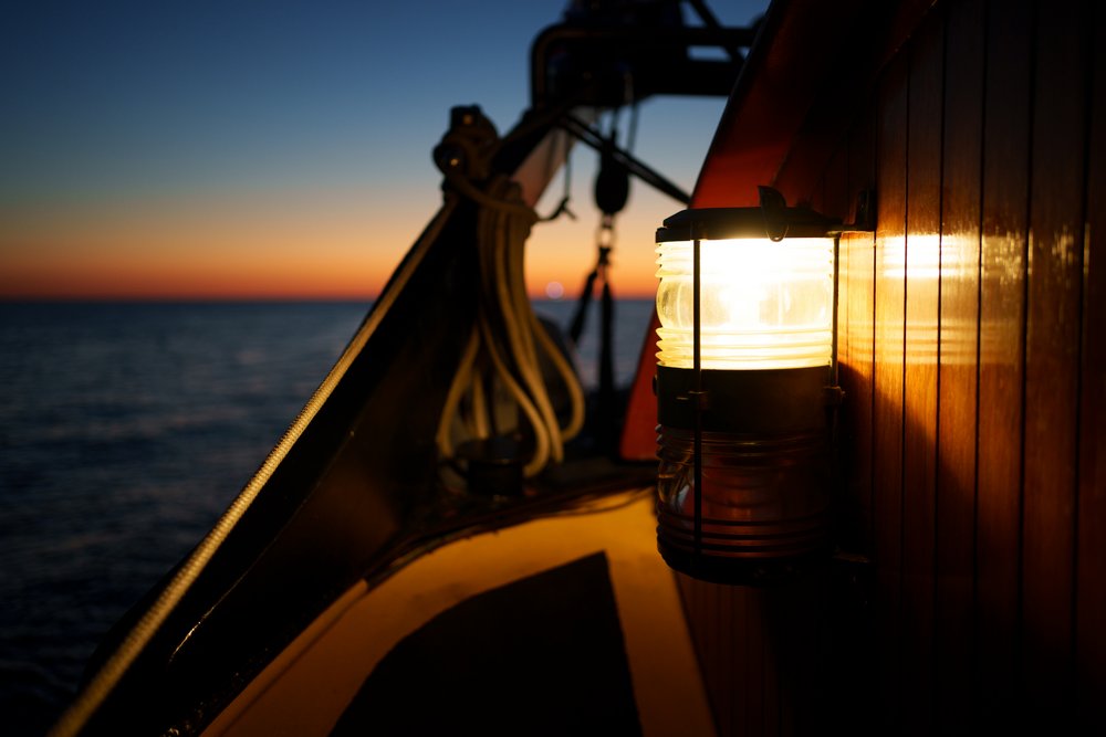 Ein nautisches Laternenlicht leuchtet an der Seite eines Schiffes bei Sonnenuntergang, während das Meer im Hintergrund in warmen und kühlen Tönen des abendlichen Himmels glitzert. Die ruhige See und das sanfte Licht der Laterne schaffen eine friedliche und beschauliche Atmosphäre auf dem Deck des Schiffes.